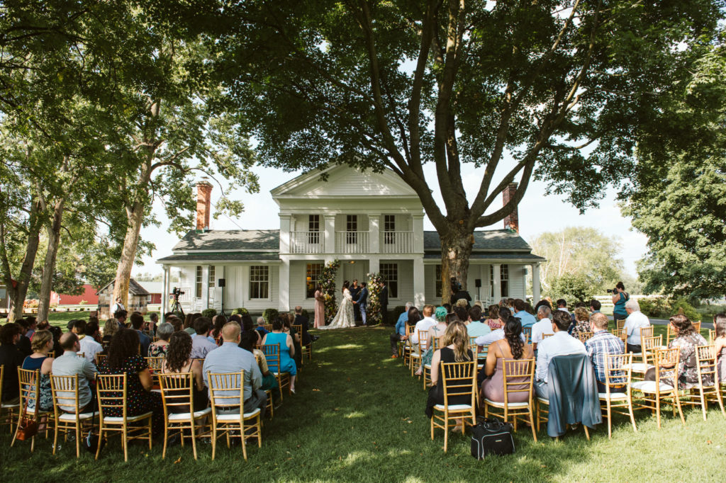 Couple getting married with guests in front of a white farmhouse and large trees